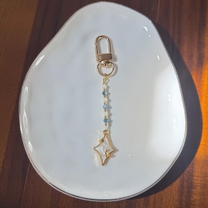 Puede incluir: Un llavero dorado con un mosquetón, una cadena de cuentas azules y blancas y un dije en forma de estrella. El dije tiene un centro blanco con un contorno dorado y un pequeño diseño de media luna.