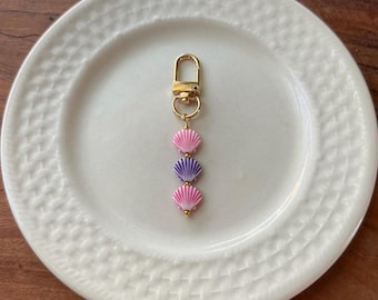 May include: A gold-coloured keychain featuring three seashell charms in pink and purple. The keychain is attached to a gold clasp. The seashells are arranged vertically on a white plate.