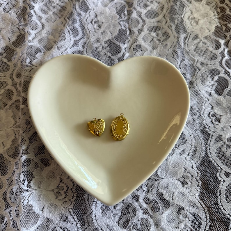 K&ouml;nnte beinhalten: Zwei kleine, goldfarbene Medaillons liegen auf einer herzf&ouml;rmigen, cremefarbenen Schale. Das eine Medaillon ist herzf&ouml;rmig mit floralem Design, das andere oval mit einem verzierten Rand. Die Schale liegt auf einer wei&szlig;en Spitzentischdecke.