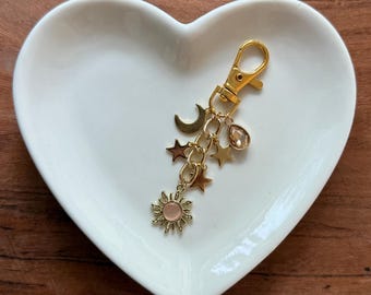 Könnte beinhalten: Goldfarbener Schlüsselanhänger mit himmlischen Anhängern, darunter ein Halbmond, Sterne und ein Sonnenanhänger mit rosa Mitte. Der Schlüsselanhänger liegt auf einer weißen herzförmigen Schale auf einer Holzoberfläche.