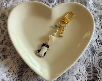 Könnte beinhalten: Ein goldfarbener Schlüsselanhänger mit einem schwarz-weißen Katzenanhänger, einem goldenen Buchstaben "E" und dekorativen Perlen. Der Schlüsselanhänger wird auf einer herzförmigen, cremefarbenen Schale präsentiert.