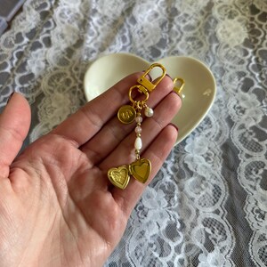 K&ouml;nnte beinhalten: Goldfarbener Schl&uuml;sselanh&auml;nger mit einem herzf&ouml;rmigen Medaillon, einem runden Anh&auml;nger mit dem Buchstaben "S" und dekorativen Perlen. Das Medaillon l&auml;sst sich &ouml;ffnen und bietet Platz f&uuml;r ein kleines Foto oder Andenken. Der Schl&uuml;sselanh&auml;nger wird in einer Hand gehalten.