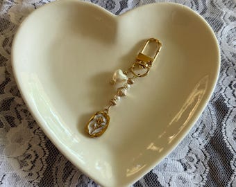 Könnte beinhalten: Ein goldfarbener Schlüsselanhänger mit einem dekorativen Anhänger. Der Anhänger hat eine goldfarbene ovale Form mit einem weißen Blumendesign. Der Schlüsselanhänger enthält auch eine herzförmige Perle und mehrere perlenartige Perlen. Der Schlüsselanhänger liegt in einer herzförmigen Schale.