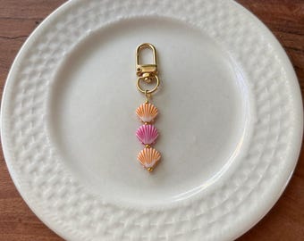 May include: A gold-coloured keyring featuring three seashell charms in shades of pink and orange. The keyring has a gold clasp, and the seashells are arranged vertically.