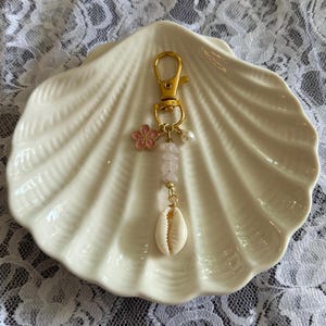 Peut inclure: Un porte-clés doré avec un mousqueton, un charm en forme de fleur rose, une chaîne de perles de quartz rose, une petite perle et un coquillage blanc. Le porte-clés est présenté sur un plat blanc en forme de coquillage.