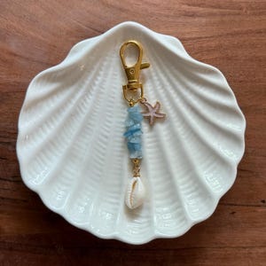 Peut inclure: Un porte-clés doré avec un fermoir mousqueton, orné de pierres bleu clair, d'un petit coquillage et d'un charm en forme d'étoile de mer rose. Le porte-clés est présenté sur un plat blanc en forme de coquillage.