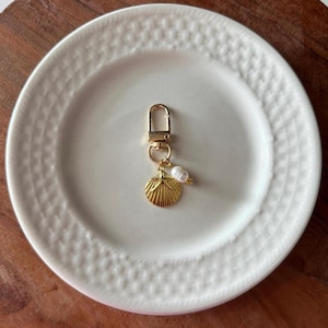 May include: A gold-colored keychain with a lobster clasp, a seashell charm, and a small pearl accent. The seashell charm has a detailed, textured design. The keychain is displayed on a white plate with a textured pattern.