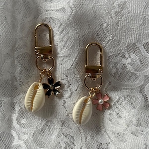 Peut inclure: Deux porte-clés dorés avec fermoirs mousquetons. Chaque porte-clés comporte un charme en forme de coquillage crème et un charme en forme de fleur. Une fleur est noire, l'autre rose avec un centre perlé. Le fond est un tissu en dentelle blanche.