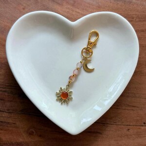 May include: A gold-colored keychain with a lobster clasp, featuring a sun charm with an orange center, a crescent moon, and pink and orange beads. The keychain is displayed on a white heart-shaped dish.