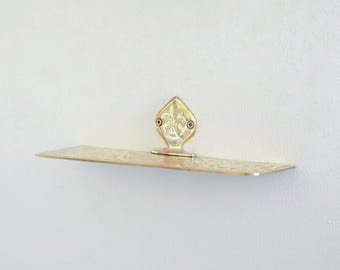 May include: A gold metal shelf with an ornate design. The shelf is mounted to a white wall and has a decorative, diamond-shaped bracket.