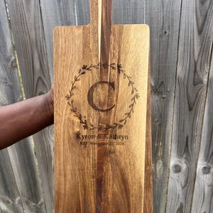 May include: A wooden cutting board with a handle, engraved with a floral wreath, the letter "C", and the text "Kyron & Kathryn Est. November 18, 2024".