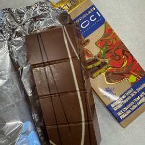 May include: A partially unwrapped chocolate bar with white and dark chocolate drizzle. The bar is next to its packaging, which has text in multiple languages. The packaging is gold and brown with red and green accents.