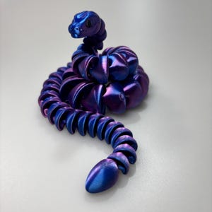 May include: A flexible, iridescent snake toy in shades of blue and purple. The toy is segmented, allowing it to bend and move. The snake's head is raised, and its body curves in a sinuous shape.