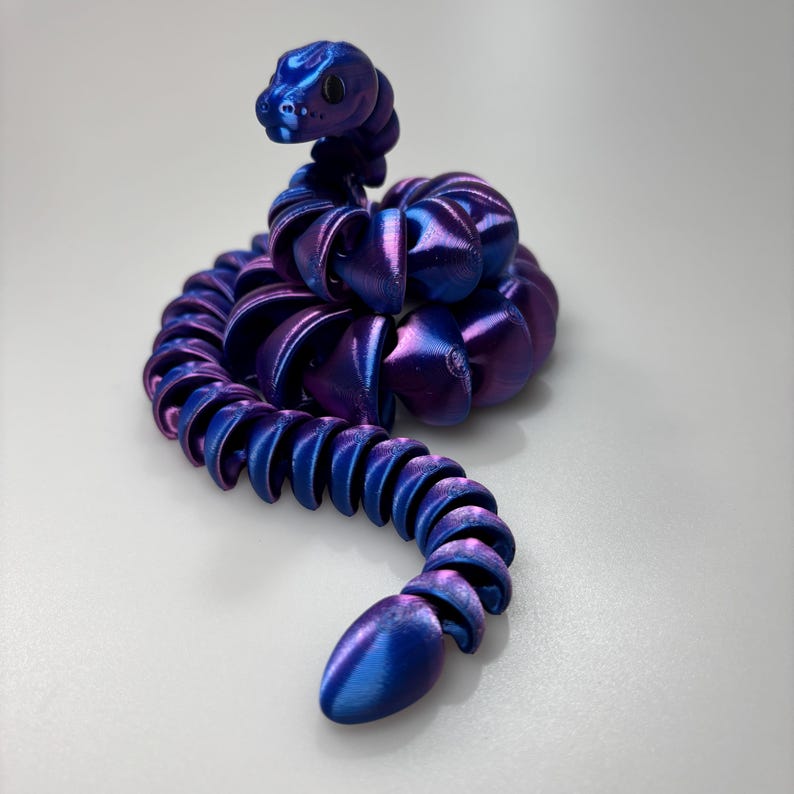 May include: A flexible, iridescent snake toy in shades of blue and purple. The toy is segmented, allowing it to bend and move. The snake's head is raised, and its body curves in a sinuous shape.