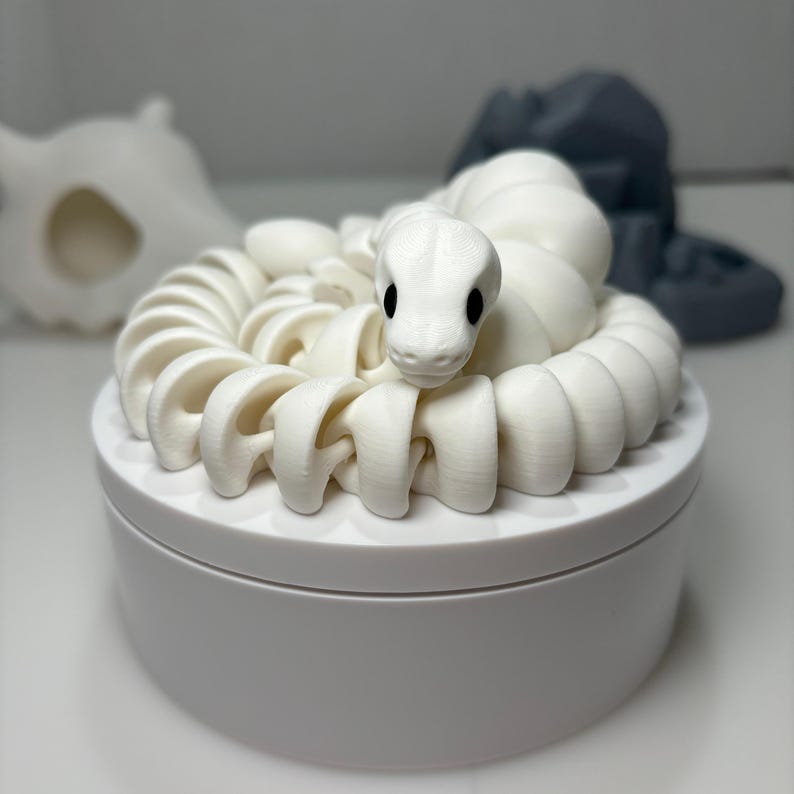 May include: A white, 3D-printed snake coiled on top of a white, round container. The snake has a detailed design with black eyes. The container is sitting on a white surface. Other blurred objects are in the background.