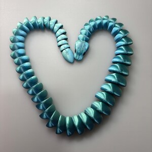 May include: A turquoise and teal snake-shaped necklace. The segmented body of the snake forms a heart shape. The necklace has a metallic sheen, with a gradient of colors from teal to turquoise.