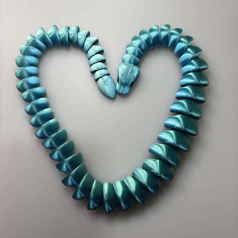 May include: A turquoise and teal snake-shaped necklace. The segmented body of the snake forms a heart shape. The necklace has a metallic sheen, with a gradient of colors from teal to turquoise.