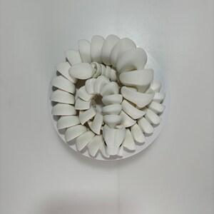 May include: A coiled, white ceramic sculpture of a snake, resting on a white circular base. The snake's body is composed of stacked, curved segments, with a detailed head and visible eyes. The artwork is set against a plain white background.