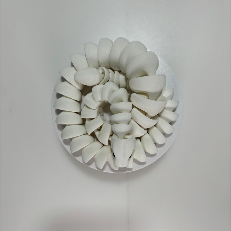 May include: A coiled, white ceramic sculpture of a snake, resting on a white circular base. The snake's body is composed of stacked, curved segments, with a detailed head and visible eyes. The artwork is set against a plain white background.
