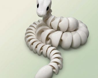 May include: A white articulated snake figurine with a rounded head and black eyes. The body is composed of connected, curved segments, creating a flexible, coiled form. The snake is posed on a light green background.