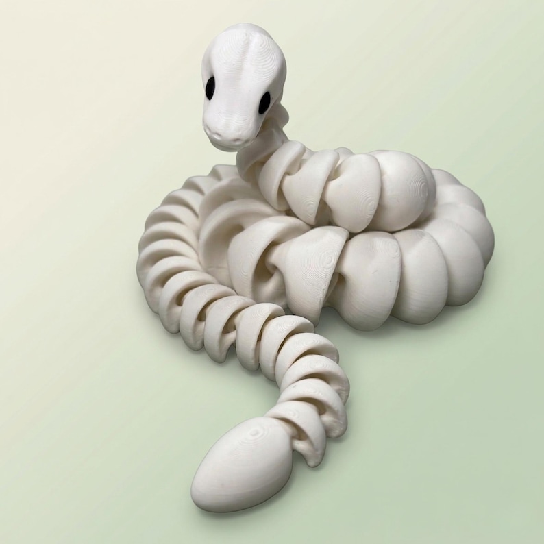 May include: A white articulated snake figurine with a rounded head and black eyes. The body is composed of connected, curved segments, creating a flexible, coiled form. The snake is posed on a light green background.