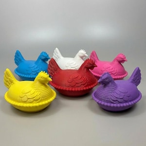 May include: A collection of colorful hen-shaped containers in blue, white, red, pink, yellow, and purple. Each container has a textured surface and a detailed design, perfect for kitchen decor or storage.