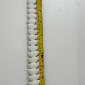 May include: A white, segmented, snake-like object is positioned vertically next to a yellow measuring tape. The tape shows measurements up to 22 inches. The object's design resembles a series of stacked, rounded bowls.