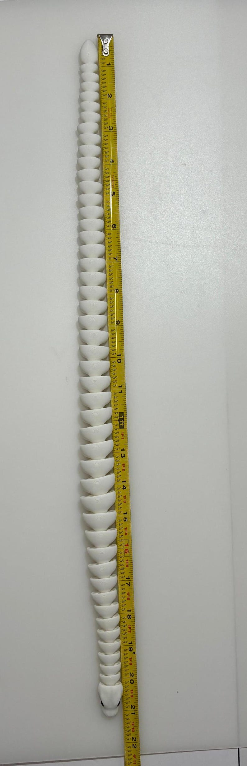 May include: A white, segmented, snake-like object is positioned vertically next to a yellow measuring tape. The tape shows measurements up to 22 inches. The object's design resembles a series of stacked, rounded bowls.