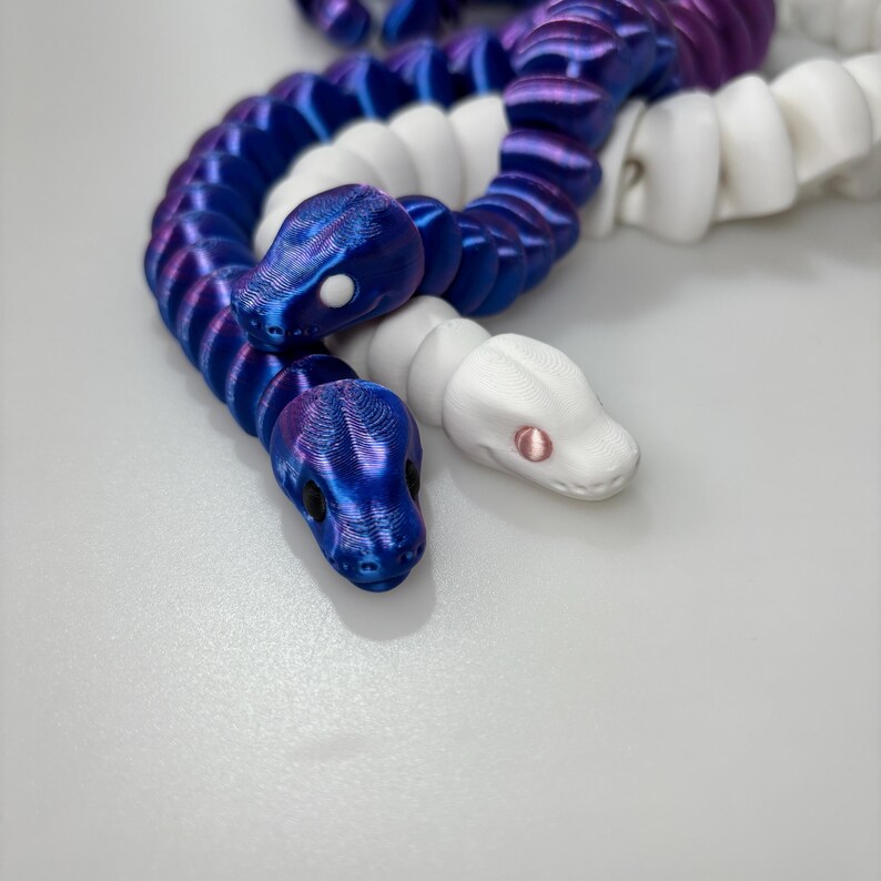 May include: Two articulated snake toys. One is a gradient of blue and purple, the other is white. The snakes have detailed scales and realistic-looking heads with small eyes. The toys are designed to be flexible and poseable.