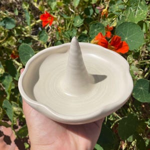 Puede incluir: Un soporte para anillos de cerámica color crema con un poste central en forma de cono y un borde festoneado. El soporte está diseñado para guardar anillos y pequeñas joyas. El fondo presenta vibrantes flores naranjas y follaje verde.