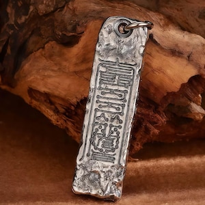 May include: A rectangular silver pendant with an engraved design and a small loop at the top. The pendant has a textured, hammered appearance and features an intricate pattern of characters. The background is a piece of wood.