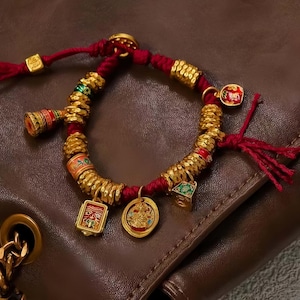May include: A gold and red bracelet with multiple charms. The bracelet features gold beads, red cord, and various gold charms with colorful enamel details. The charms include a square face, a round medallion, and a small green cube.