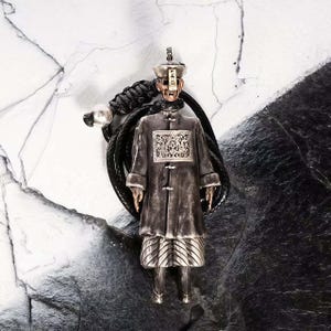 Könnte beinhalten: Ein silberner Anhänger in Form eines chinesischen Jiangshi, oder Hüpfvampirs, mit detailliertem Mantel und Hut. Der Anhänger ist an einer schwarzen Kordel mit einer silbernen Perle befestigt.