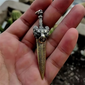 Op de afbeelding: Een gedetailleerde hanger in de vorm van een klein zwaard. De hanger heeft een decoratief handvat met een gedetailleerd dierenkopontwerp en een lemmet met gegraveerde patronen. Het metaal heeft een zilveren en bronzen kleur.