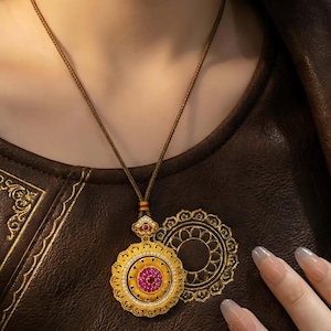 May include: A gold pendant necklace with a circular design featuring a central pink gemstone surrounded by smaller yellow stones and clear crystals. The pendant hangs from a brown cord. The necklace is displayed on a brown leather surface.