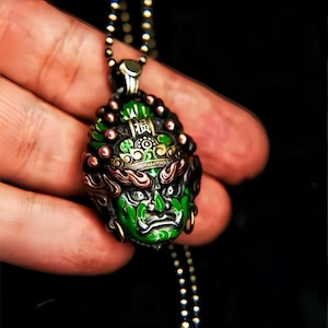 Può includere: Un ciondolo d'argento dettagliato con un design a volto verde e nero, con dettagli intricati e una piccola iscrizione. Il ciondolo è appeso a una catena di perline. Il volto ha un'espressione feroce ed è accentuato da dettagli rossi e dorati.