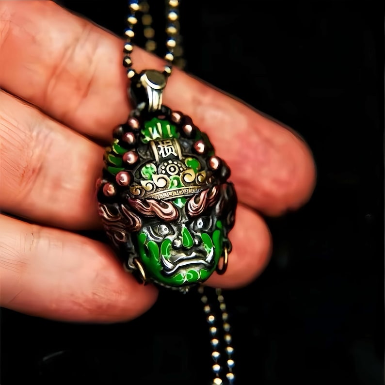 May include: A detailed silver pendant with a green and black face design, featuring intricate details and a small inscription. The pendant hangs from a beaded chain. The face has a fierce expression and is accented with red and gold details.