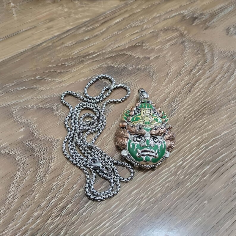May include: A silver-toned necklace with a detailed pendant featuring a green, gold, and bronze-colored mask. The mask has intricate facial features and is attached to a chain with a lobster clasp.