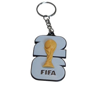May include: A keychain with a 3D-printed gold-coloured FIFA World Cup trophy. The trophy is set against a white background with the number "23" and the word "FIFA" in brown. The keychain has a silver ring and chain.
