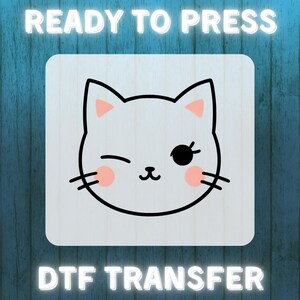以下が含まれることがあります： 白い猫が薄い青色の背景に描かれており、黒い眼帯とピンクの鼻をしています。猫は片目を閉じ、ウィンクをしています。猫の上に「READY TO PRESS」という文字が、猫の下に「DTF TRANSFER」という文字が書かれています。