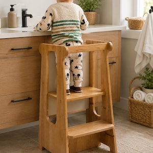 May include: A light brown wooden learning tower with two steps and a safety rail, designed to provide elevation for a child to reach a bathroom counter. The structure features a sturdy frame with smooth edges and a natural wood grain finish.