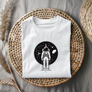 Pode incluir: Camiseta branca com uma ilustração em preto e branco de um astronauta no espaço. O astronauta está vestindo um traje espacial e um capacete. A ilustração inclui um foguete, planetas e estrelas.