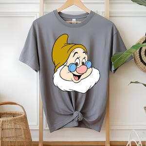 以下が含まれることがあります： 黄色い帽子、青いメガネ、白いひげのキャラクターの漫画のイメージがプリントされたグレーのTシャツ。キャラクターはピンクの鼻をしていて、笑顔です。Tシャツはウエストで結ばれています。