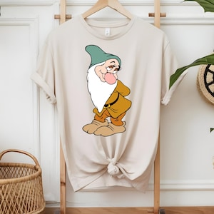 以下が含まれることがあります： ベージュ色のTシャツ。漫画の小人のイラストが描かれています。小人は白いひげ、緑の帽子、茶色の衣装をしています。Tシャツは木製のラックに展示されています。