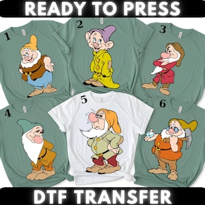 以下が含まれることがあります： 7人の小人の漫画の画像がプリントされた6枚のTシャツ。シャツは緑と白など様々な色があります。「READY TO PRESS」と「DTF TRANSFER」の文字も表示されています。
