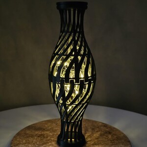 Könnte beinhalten: Schwarze dekorative Vase mit Gittermuster, beleuchtet von warmweißen Lichterketten. Die Vase besteht aus dunklem Material und steht auf einer runden Korkoberfläche. Das Gesamtdesign ist modern und elegant.