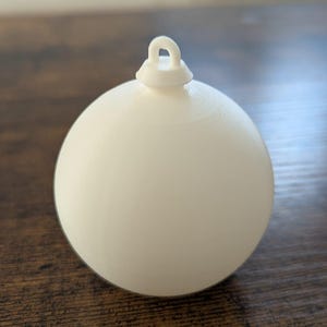 Könnte beinhalten: Ein weißer, kugelförmiger Weihnachtsschmuck mit einer Schlaufe zum Aufhängen. Der Schmuck hat eine glatte, matte Oberfläche und liegt auf einer dunklen Holzoberfläche. Der Schmuck ist ein schlichtes, klassisches Design.
