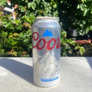 以下が含まれることがあります： 赤い「Coors」のロゴと山のグラフィックが入った、銀色のCoorsビール缶。「COLD AS THE ROCKIES」と「WHEN THE MOUNTAIN TURNS BLUE IT'S AS COLD AS THE ROCKIES」という文字が入っています。ロゴの上には青い三角形があります。