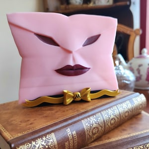 Puede incluir: Una escultura rosa con forma de rostro con labios y formas de ojos de color burdeos oscuro y un detalle de lazo dorado. La escultura descansa sobre una pila de libros antiguos con letras doradas. Un juego de té decorativo está en el fondo.