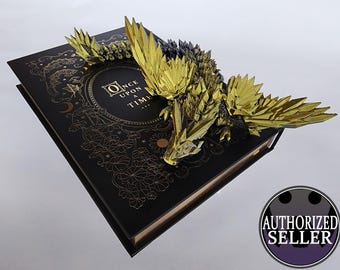 Puede incluir: Un libro negro con detalles en pan de oro y una escultura de dragón dorada en la parte superior. La portada del libro tiene el texto "Once Upon a Time..."